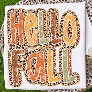 Puede incluir: Una camiseta blanca con el texto HELLO FALL en letras grandes y gruesas. Cada letra está rellena con un patrón de calabazas y hojas, delineada con un borde de estampado de leopardo. Los colores incluyen tonos naranja, marrón y verde salvia.