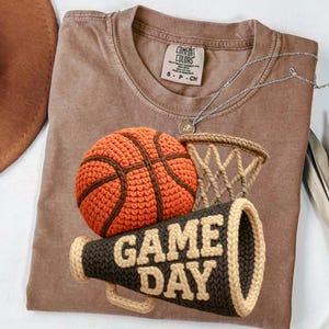K&ouml;nnte beinhalten: Braunes T-Shirt mit einem geh&auml;kelten Basketball, Netz und Megaphon mit der Aufschrift "GAME DAY". Der Basketball ist orange mit braunen Linien. Das Megaphon ist dunkelbraun mit beigen Verzierungen und Schrift. Eine silberne Halskette liegt auf dem Shirt.