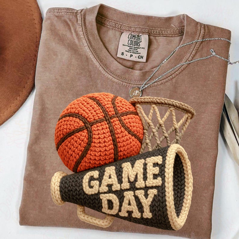 K&ouml;nnte beinhalten: Braunes T-Shirt mit einem geh&auml;kelten Basketball, Netz und Megaphon mit der Aufschrift "GAME DAY". Der Basketball ist orange mit braunen Linien. Das Megaphon ist dunkelbraun mit beigen Verzierungen und Schrift. Eine silberne Halskette liegt auf dem Shirt.
