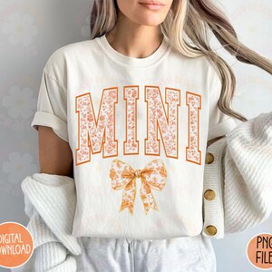 May include: White t-shirt with the word "MINI" in orange floral letters and a floral bow.