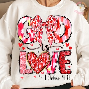 May include: White sweatshirt with a pink and red heart design. The text "God is Love" is written in a bold font with a red heart in the middle of the word "Love". The text "1 John 4:8" is written below the design.