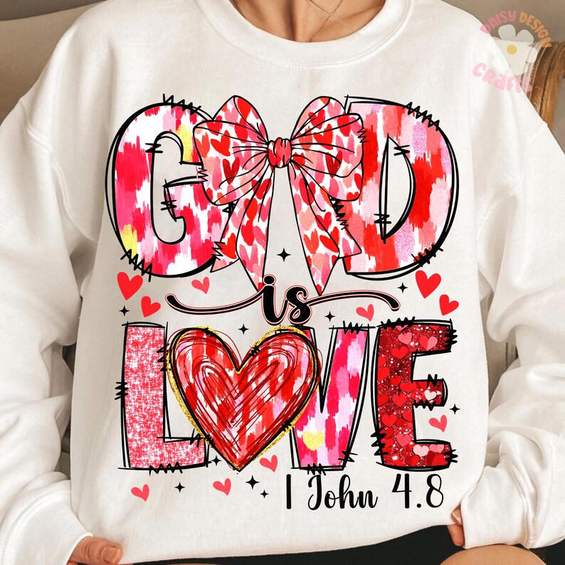 May include: White sweatshirt with a pink and red heart design. The text "God is Love" is written in a bold font with a red heart in the middle of the word "Love". The text "1 John 4:8" is written below the design.