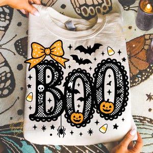 Puede incluir: Una sudadera crema con un dise&ntilde;o tem&aacute;tico de Halloween. La palabra "BOO" est&aacute; escrita en letras negras ornamentadas, con un lazo, murci&eacute;lagos, calabazas, estrellas y caramelos. El dise&ntilde;o es sostenido por una persona.