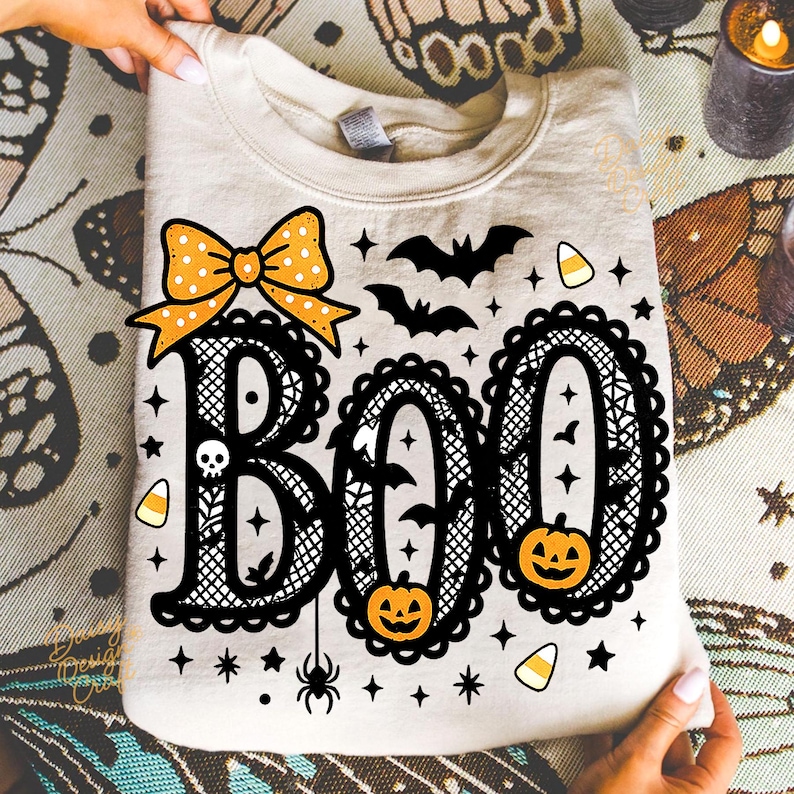 Puede incluir: Una sudadera crema con un dise&ntilde;o tem&aacute;tico de Halloween. La palabra "BOO" est&aacute; escrita en letras negras ornamentadas, con un lazo, murci&eacute;lagos, calabazas, estrellas y caramelos. El dise&ntilde;o es sostenido por una persona.