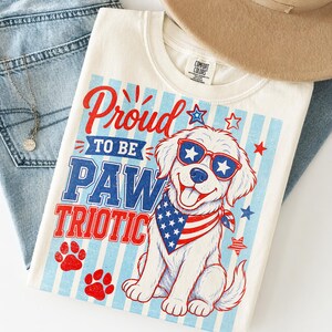 May include: Cream-colored t-shirt with a patriotic dog graphic. The design features a smiling dog wearing sunglasses and a bandana with the American flag. Text reads "Proud to be Paw Triotic."