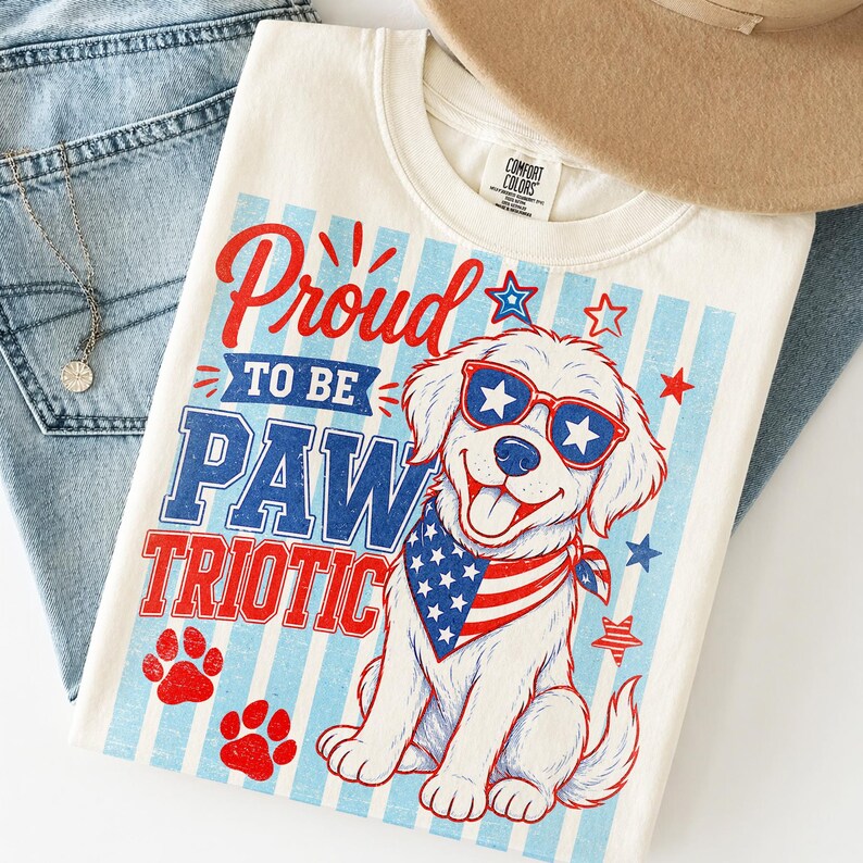 May include: Cream-colored t-shirt with a patriotic dog graphic. The design features a smiling dog wearing sunglasses and a bandana with the American flag. Text reads "Proud to be Paw Triotic."
