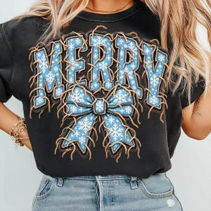 May include: A black short-sleeved t-shirt featuring the word MERRY in large blue letters with a white snowflake pattern. The letters and a large blue bow graphic below are outlined with a brown, frayed, rope-like texture effect.