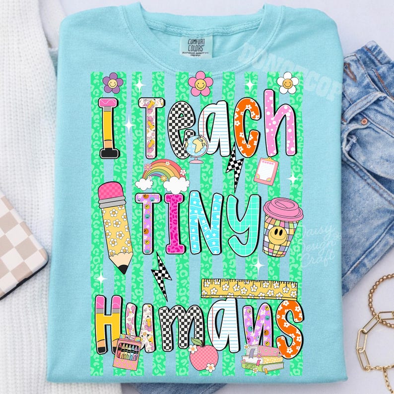 Puede incluir: Una camiseta azul claro con un gr&aacute;fico colorido y el texto I TEACH TINY HUMANS. El dise&ntilde;o incluye un l&aacute;piz, un arco&iacute;ris, un globo terr&aacute;queo, una taza de caf&eacute;, una regla y libros, sobre un fondo de rayas verticales con estampado de leopardo verde.