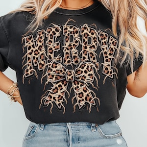 May include: A black crewneck sweatshirt featuring the word MERRY in large, distressed-edge letters with a leopard print pattern. Below the text is a large, matching leopard print bow with frayed edges, creating a textured appearance.