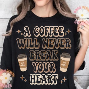 Puede incluir: Camiseta negra con un gr&aacute;fico de estilo retro que dice "Un caf&eacute; nunca romper&aacute; tu coraz&oacute;n". El gr&aacute;fico presenta dos tazas de caf&eacute; helado.