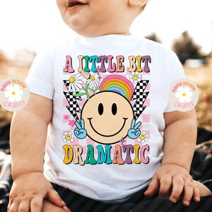 Op de afbeelding: Een wit T-shirt met een regenboog, een smiley en de tekst "A little bit dramatic" in kleurrijke letters.
