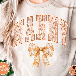 May include: A white sweatshirt with the word "NANNY" in orange letters with a floral pattern. The letters are outlined in orange. A large orange bow with a floral pattern is below the word.