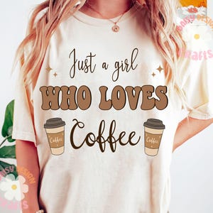Puede incluir: Una camiseta blanca con el texto "Just a girl who loves coffee" en letras marrones. Dos tazas de caf&eacute; marrones con la palabra "coffee" en ellas est&aacute;n a cada lado del texto.