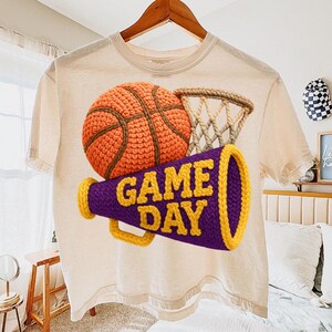 K&ouml;nnte beinhalten: Cremefarbenes T-Shirt mit einem geh&auml;kelten Basketball, Netz und Megaphon. Das Megaphon ist lila mit gelben Verzierungen und den Worten "GAME DAY" in Gelb. Der Basketball ist orange und braun.