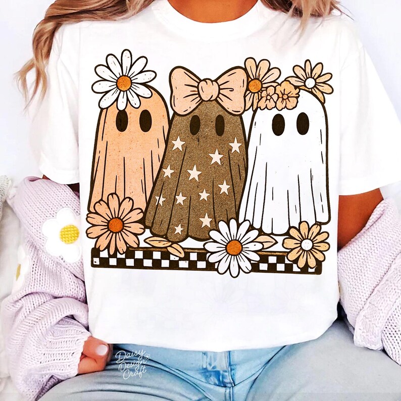 May include: White t-shirt featuring a graphic of three cartoon ghosts with daisy accents. The ghosts are in shades of peach, brown with white stars, and white. The design includes a black and white checkered border and the text "Daisy Design Craft".