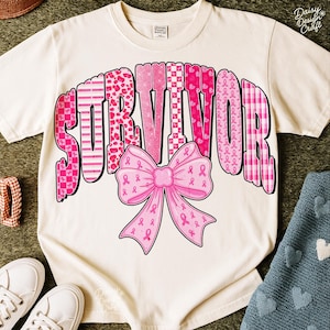 May include: Cream-colored t-shirt with the word "SURVIVOR" in pink, patterned letters. A pink bow with pink ribbons is centered below the word. The shirt is a casual style, suitable for everyday wear.