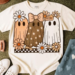 May include: Cream-colored t-shirt featuring a whimsical design of three ghost figures adorned with daisies and bows. The ghosts are in shades of peach, brown, and white, with star and daisy accents. A black and white checkered border is at the bottom.