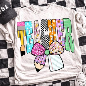 Puede incluir: Una camiseta de color crema con la palabra TEACHER en letras grandes con varios patrones. El dise&ntilde;o incluye un pincel, un tablero de ajedrez en blanco y negro, estampados florales, caras sonrientes y un gran lazo rosa con un l&aacute;piz y un cuaderno.