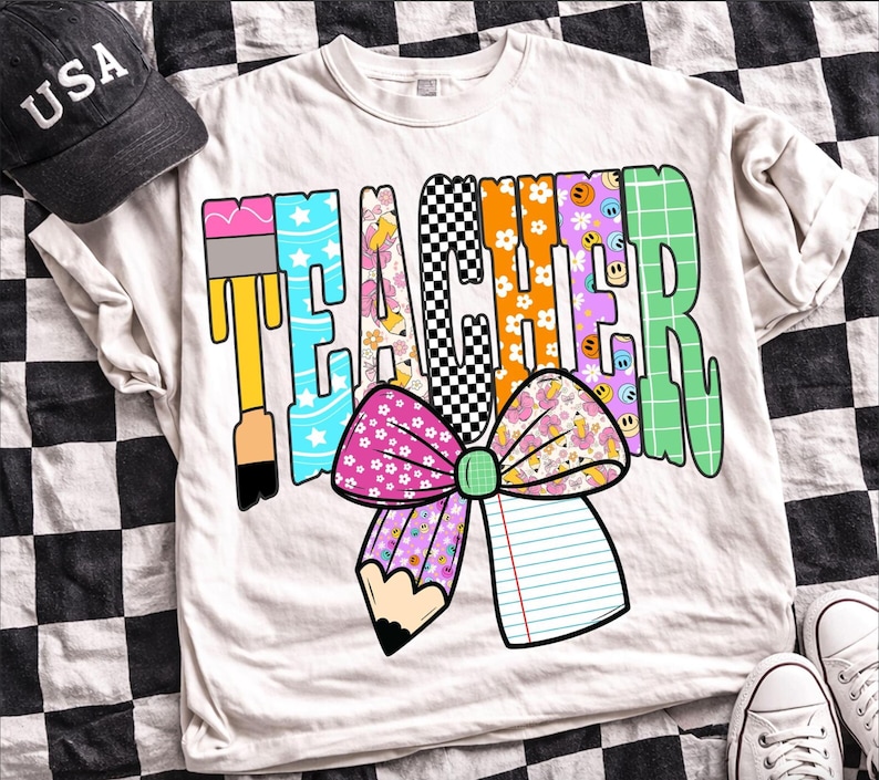Puede incluir: Una camiseta de color crema con la palabra TEACHER en letras grandes con varios patrones. El dise&ntilde;o incluye un pincel, un tablero de ajedrez en blanco y negro, estampados florales, caras sonrientes y un gran lazo rosa con un l&aacute;piz y un cuaderno.