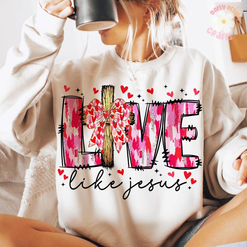May include: White sweatshirt with the word "LIVE" in pink and red, featuring a bow and cross design. The phrase "like jesus" is written below in black script, with red heart accents. A casual and inspirational design.