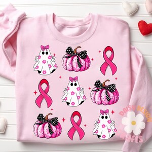 May include: Pink sweatshirt with a pattern of pink pumpkins, ghosts, and pink ribbons. The design is a cute and whimsical take on Halloween and breast cancer awareness.