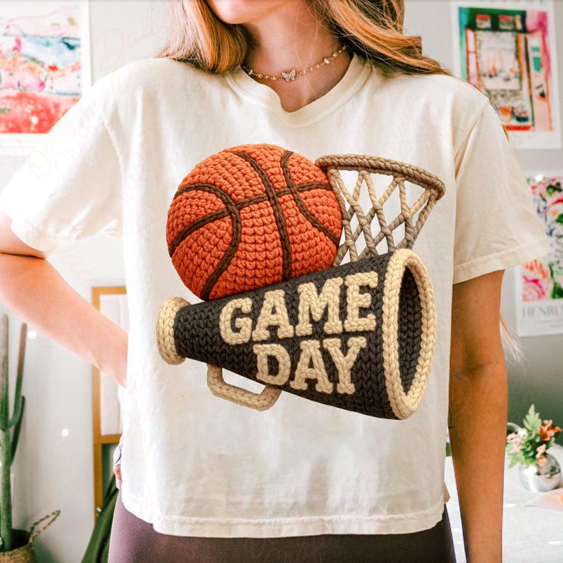 K&ouml;nnte beinhalten: Cremefarbenes T-Shirt mit einem geh&auml;kelten Basketball, Korb und Megaphon. Der Basketball ist orange mit braunen Linien, der Korb ist beige und das Megaphon ist dunkelbraun mit "GAME DAY" in beigen Buchstaben.