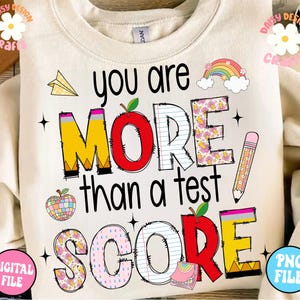 Può includere: Una felpa beige con il testo "You are more than a test score" in nero. Le lettere sono decorate con motivi colorati, tra cui matite, mele e arcobaleni.