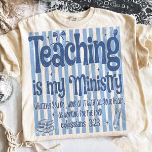 Puede incluir: Una camiseta de color crema con un fondo de rayas verticales azules y el texto "Teaching is my Ministry" en una fuente grande y negrita. Debajo, se lee "Whatever you do, work at it with all your heart, as working for the Lord, Colossians 3:23", acompa&ntilde;ado de una pila de libros y un gr&aacute;fico de un l&aacute;piz.