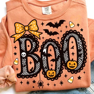 Puede incluir: Sudadera color melocot&oacute;n con un dise&ntilde;o tem&aacute;tico de Halloween. La palabra "BOO" est&aacute; escrita en una fuente decorativa con un lazo, murci&eacute;lagos, calabazas, estrellas y una ara&ntilde;a. La sudadera es de material suave.