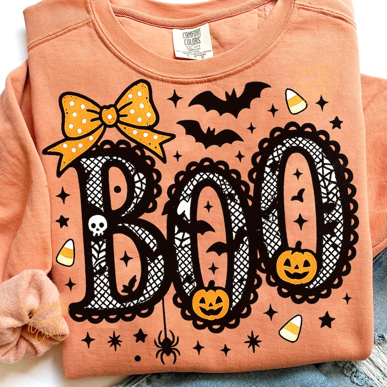 Puede incluir: Sudadera color melocot&oacute;n con un dise&ntilde;o tem&aacute;tico de Halloween. La palabra "BOO" est&aacute; escrita en una fuente decorativa con un lazo, murci&eacute;lagos, calabazas, estrellas y una ara&ntilde;a. La sudadera es de material suave.