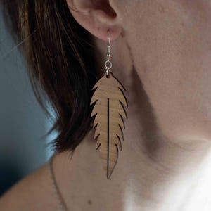 Könnte beinhalten: Ein Paar Ohrringe aus Holz in Form von Federn. Die Ohrringe sind hellbraun und haben eine natürliche Holzmaserung. Sie hängen an silbernen Haken.