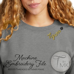 May include: Gray sweatshirt with yellow embroidered text "Tigers" and a paw print. The text "Machine Embroidery File" and "By MK" are also visible.
