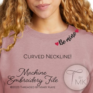 May include: A pink sweatshirt with a curved neckline and the text "Be mine" embroidered in black with red hearts. The text "Machine Embroidery File" is written in black cursive with the copyright symbol and the text "©2023 Threaded by Mary Kaye" below. A logo with a needle and thread is in the bottom right corner with the text "BY MK" in black.
