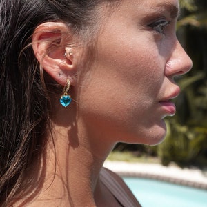 Pode incluir: Uma mulher com cabelo comprido e escuro usando um brinco em forma de coração azul com um aro dourado.