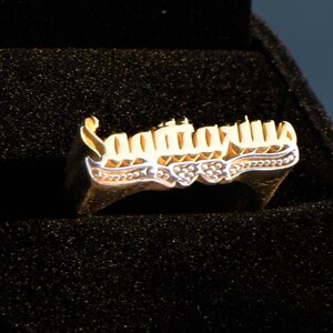 May include: A gold ring with the word "Lazarus" engraved in cursive lettering. The ring is in a black velvet ring box.