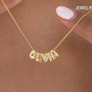 May include: Gold necklace with the name "Olivia" spelled out in individual letters. The letters are connected by a delicate chain.