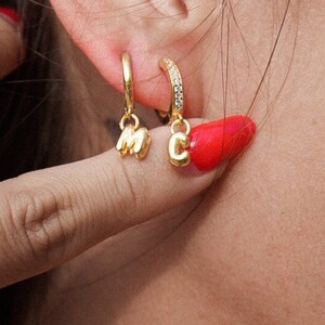 May include: A close-up of a person's ear with two gold hoop earrings. The earrings have small charms hanging from them, one is a letter 'M' and the other is a letter 'C'.