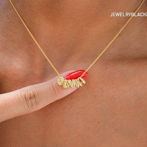 May include: Gold necklace with the name "Olivia" spelled out in individual letter charms. The necklace is worn by a person with dark skin.