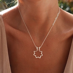 May include: A silver necklace with a geometric pendant in the shape of an eight-pointed star. The necklace is worn on a person with fair skin, wearing a beige tank top. The text "jewelryblackpearl" is written on the necklace chain.