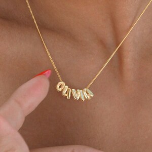 May include: Gold necklace with the name "Olivia" spelled out in individual letters. The letters are connected by a delicate chain.