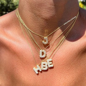 Op de afbeelding: Een close-up van een persoon die een gelaagde gouden ketting draagt met diamant-geïncruste initiaal hangers. De letters zijn "J", "D", "H", "B", en "E".