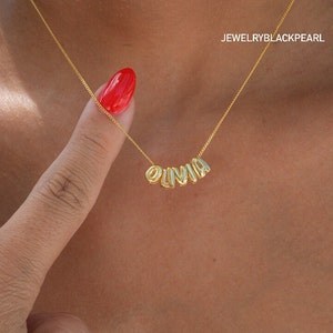 May include: A gold chain necklace with the name "OLIVIA" spelled out in gold letters. The necklace is worn by a person with a red nail polish.