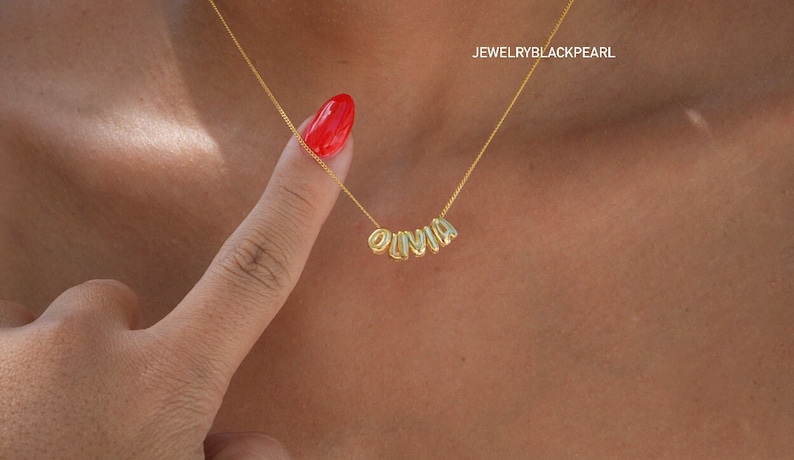 May include: A gold chain necklace with the name "OLIVIA" spelled out in gold letters. The necklace is worn by a person with a red nail polish.