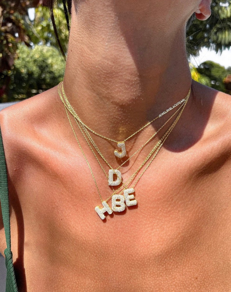 Puede incluir: Primer plano de una persona que lleva un collar de cadena de oro en capas con colgantes de iniciales engastados con diamantes. Las letras son "J", "D", "H", "B" y "E".