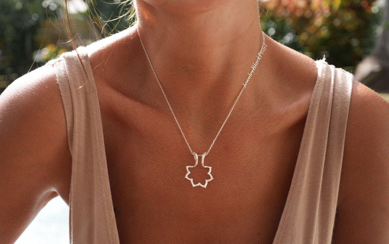 May include: A silver necklace with a geometric pendant in the shape of an eight-pointed star. The necklace is worn on a person with fair skin, wearing a beige tank top. The text "jewelryblackpearl" is written on the necklace chain.
