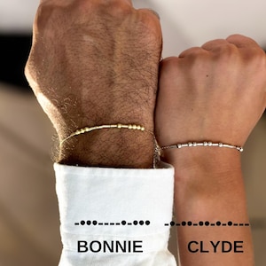 May include: Two bracelets with beads, one gold and one silver, on two wrists. The bracelets have the names "BONNIE" and "CLYDE" written below them.