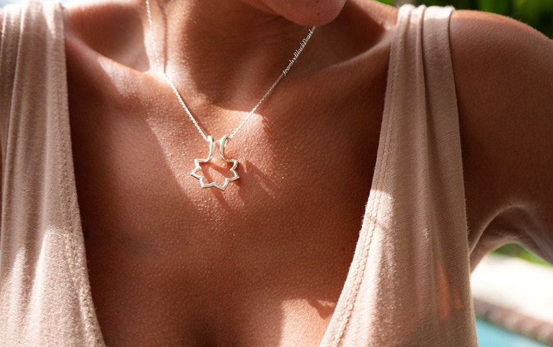 May include: A silver necklace with a pendant in the shape of an eight-pointed star. The necklace is worn against a person's skin, with a beige top visible in the background. The pendant is a delicate, openwork design.