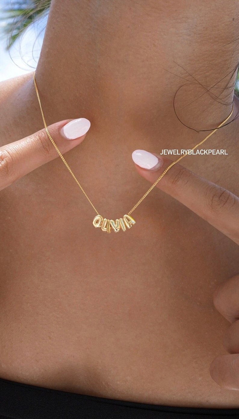 May include: Gold necklace with the name "Olivia" spelled out in individual letters. The letters are connected by a delicate chain.