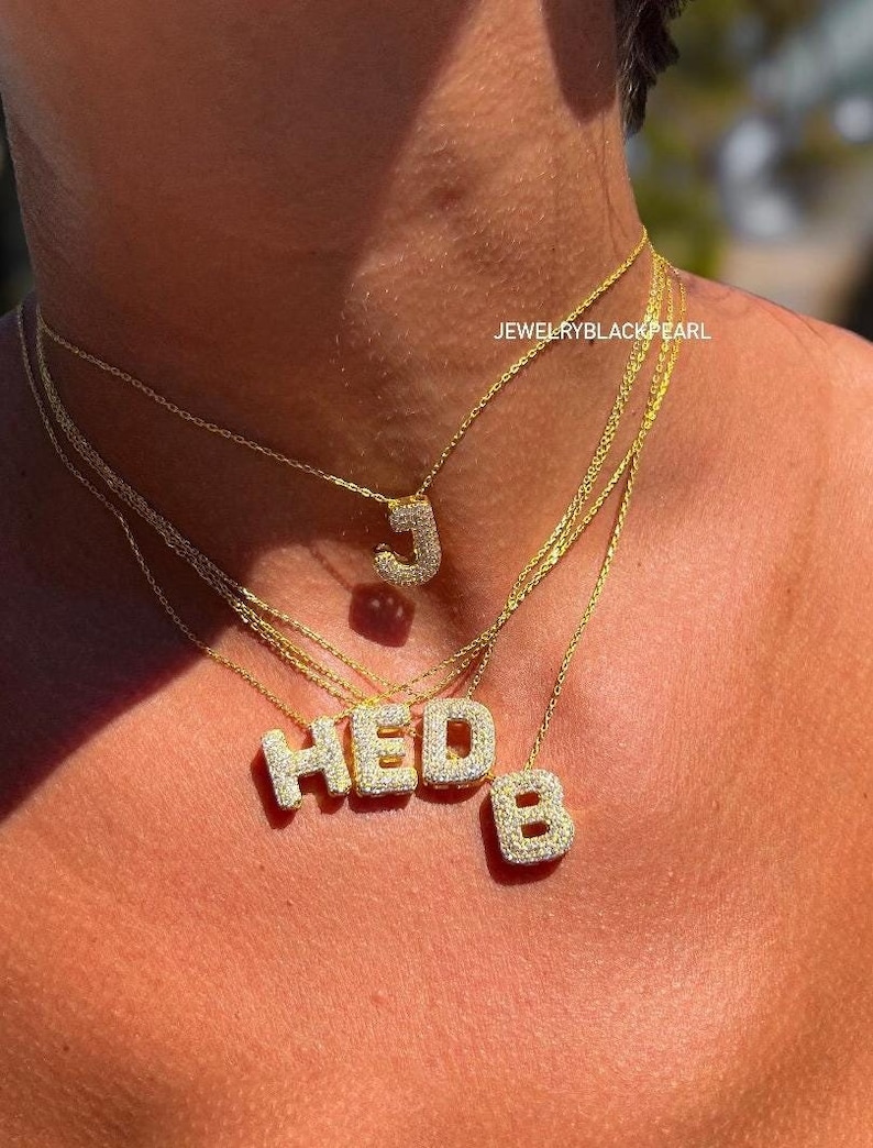 Puede incluir: Un collar de cadena de oro con m&uacute;ltiples colgantes, cada uno con una letra engastada con piedras de circonita c&uacute;bica brillantes. Las letras deletrean "HEDB". El collar lo lleva una persona, y el texto "JEWELRYBLACKPEARL" es visible en la imagen.