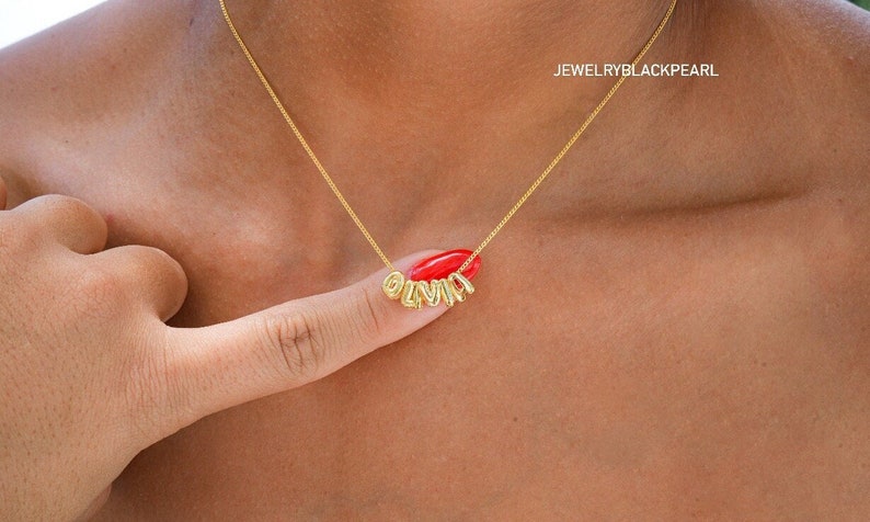 May include: Gold necklace with the name "Olivia" spelled out in individual letter charms. The necklace is worn by a person with dark skin.
